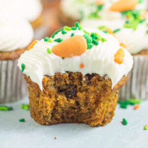 Carrot cake cupcake with a bite taken out. It is topped with cream cheese frosting, a buttercream carrot, and green jimmie sprinkles.