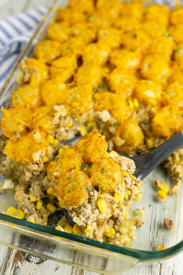 A serving of cheesy tater tot casserole on a spatula in a 9x13 baking dish on a rustic white wood background. 