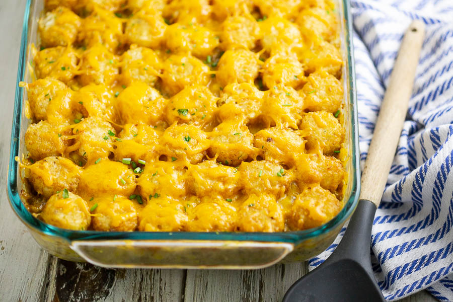 Cheesy Tater Tot Casserole next to a wooden spoon and a white and blue striped linen on a rustic white wood background.