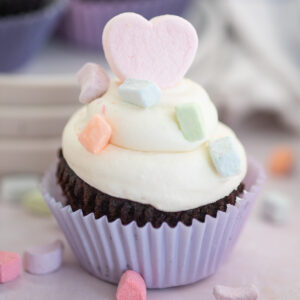 A close up of a chocolate cupcake topped with piped marshmallow buttercream frosting, a pink marshmallow heart, and colorful freeze-dried marshmallows.