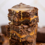 A close up of a stack of caramel brownies with a bite missing from the top one with caramel drizzled on top dripping down.