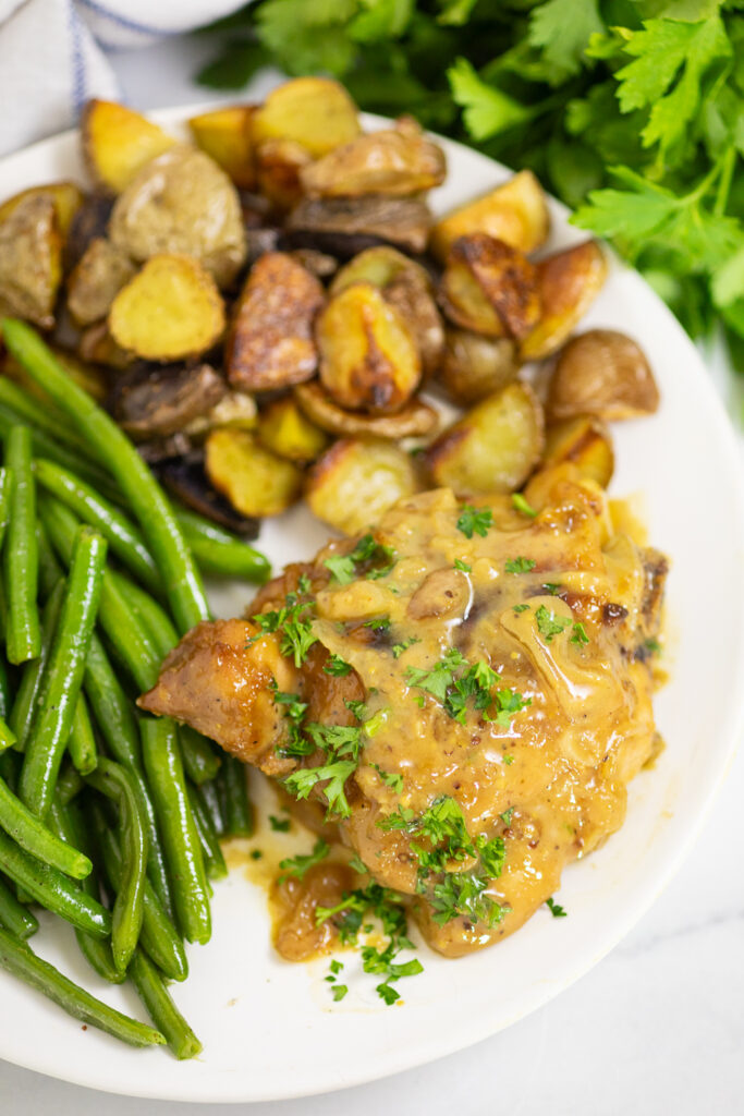 Honey mustard chicken thigh on a white plate topped with creamy honey mustard sauce and fresh parsley, next to sauteed green beans and roasted baby potatoes.