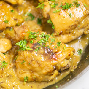 A chicken thigh, surrounded by more chicken thighs, in a cast iron skillet, smothered in creamy tangy honey mustard sauce and topped with fresh parsley.