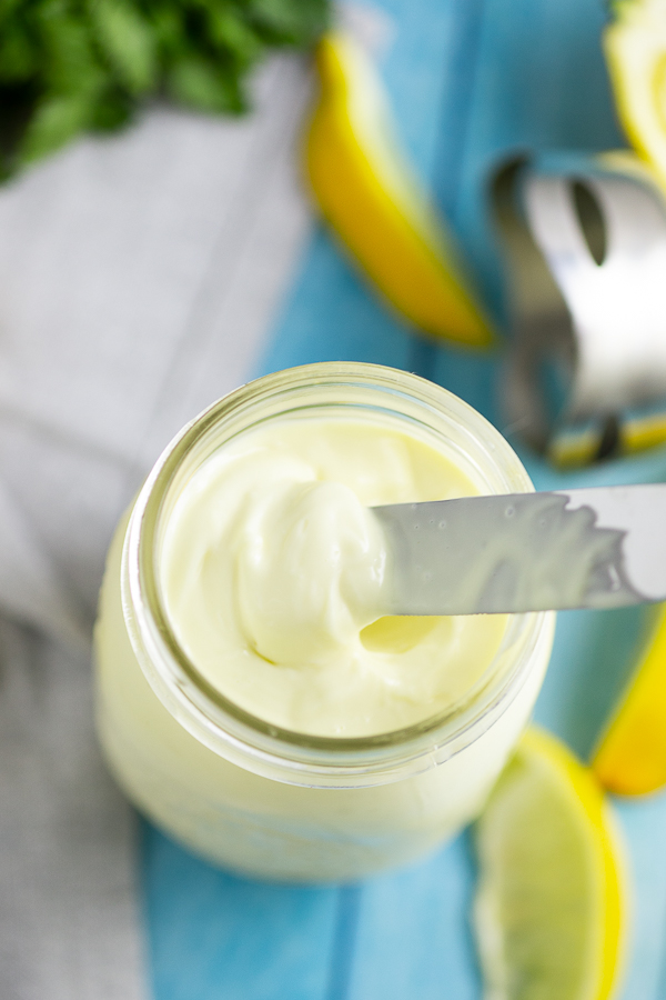 Homemade mayo in a mason jar with a butter knife scooping some out, with lemons and an immersion blender in the background