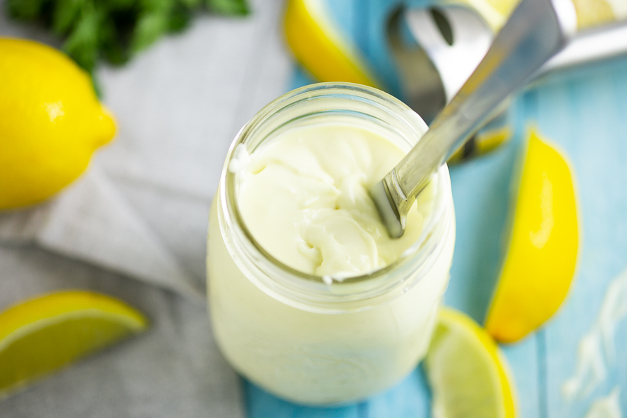 Homemade mayo with a knife in a mason jar on a bright blue background with lemon wedges.