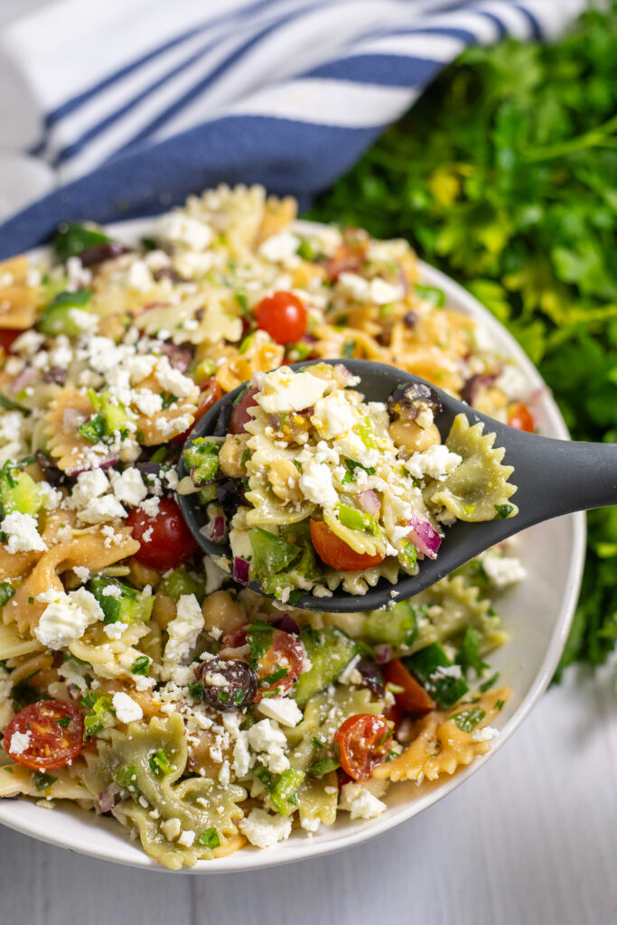 A spoon scooping up easy feta pasta salad with a blue striped linen napkin a bunch of fresh parsley behind.
