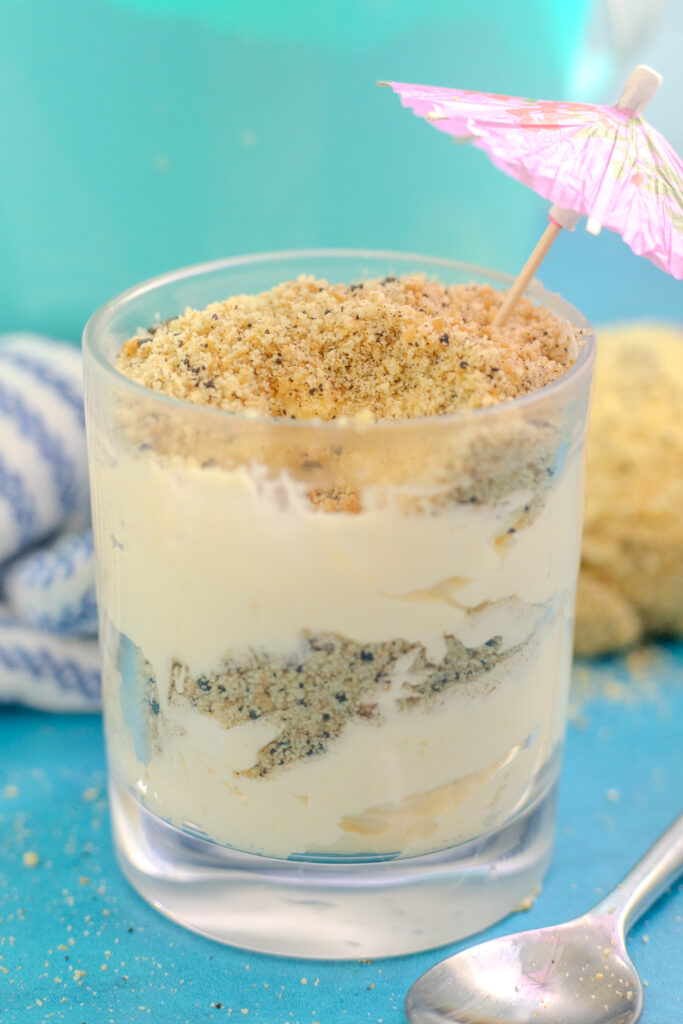 Sand pudding in a small glass with layers of pudding and cookie "sand" and a toothpick umbrella on top. The glass is on a bright blue background next to a metal spoon with a linen behind.