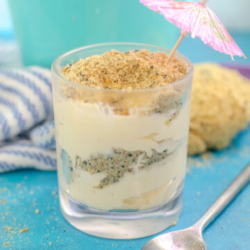 Sand pudding in a small glass with layers of pudding and cookie "sand" and a toothpick umbrella on top. The glass is on a bright blue background next to a metal spoon with a linen behind.