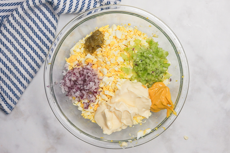 Egg salad ingredients in a glass bowl on a white marble counter