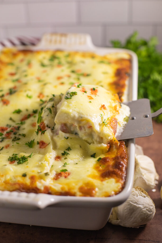 Piece of chicken alfredo lasagna being lifted out of a full large casserole dish of lasagna topped with fresh parsley and diced tomatoes with a bunch of parsley in the background