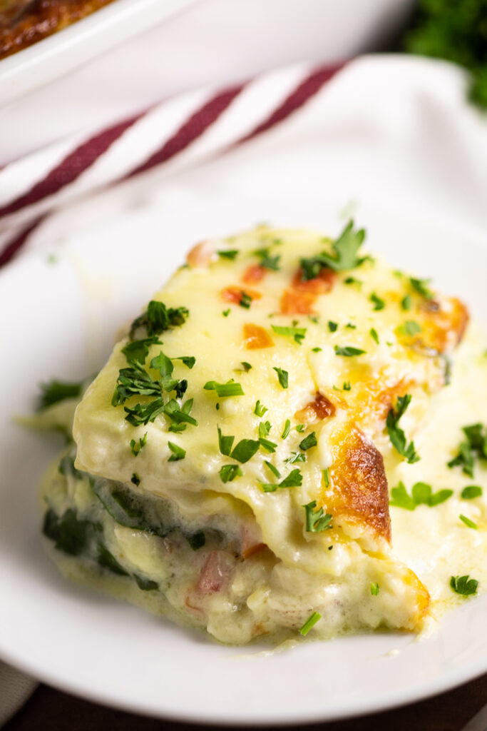 Overhead view of a serving of chicken alfredo lasagna on a small white plate topped with fresh chopped parsley with a white linen with red stripes behind