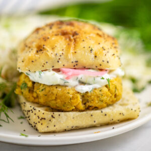 A salmon burger on a poppy seed bun topped with tzatziki and pickled red onions with coleslaw on the plate behind.