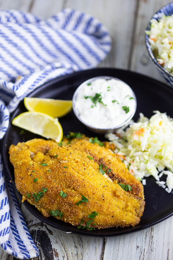 Single southern pan fried catfish filet on a plate with lemon wedges, tartar sauce, and coleslaw.