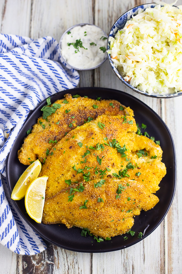 Overhead view of 3 pan fried catfish filets on a plate with lemon wedges with bowls of coleslaw and tartar sauce on the side