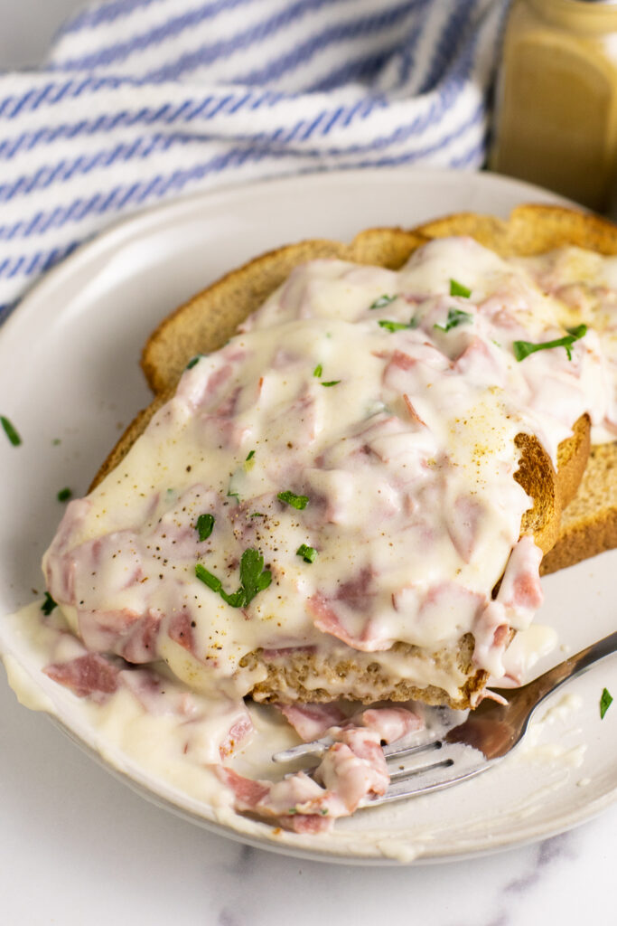 Three slices of toast smothered in creamed chipped beef and topped with black pepper and freshly chopped parsley. A fork is sitting on the small plate with the toast with a small bite taken from the toast.
