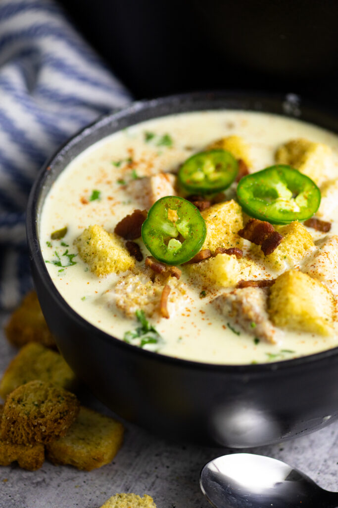 Close up of jalapeno popper soup in a black matte bowl topped with croutons, crumbled bacon, and fresh sliced jalapenos
