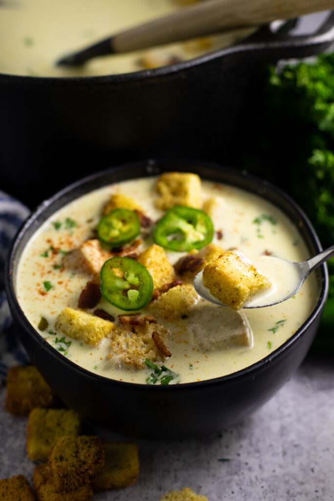 A spoon taking a bite out of a bowl of jalapeno popper soup in front of a large Dutch oven