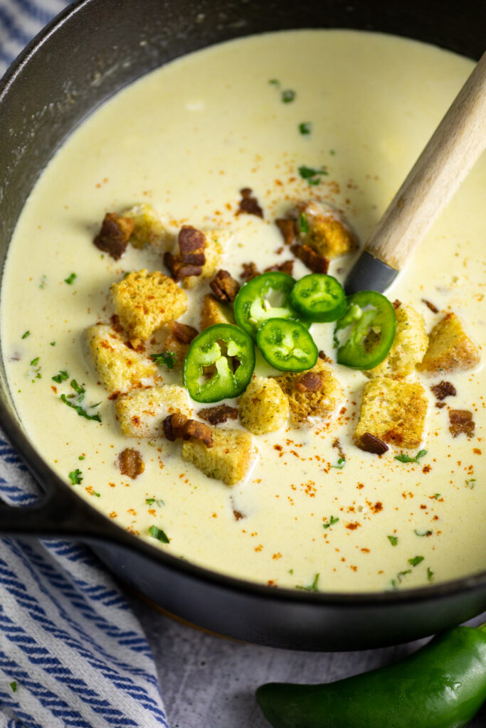 Large cast iron Dutch oven full of creamy jalapeno soup topped with croutons, crumbled bacon, and fresh jalapeno slices with a wooden ladle in the middle