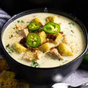 Black matte bowl full of jalapeno popper soup topped with fresh jalapeno slices and croutons next to a spoon and cloth napkin