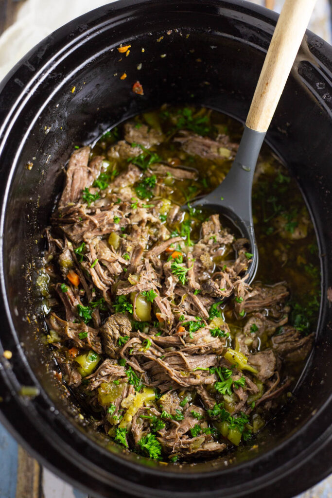 Crockpot Italian Beef in a slow cooker topped with fresh chopped parsley with a wooden spoon sticking in it.