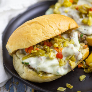 Italian Beef Sandwich topped with provolone and giardiniera on a black plate with a second sandwich sitting behind.