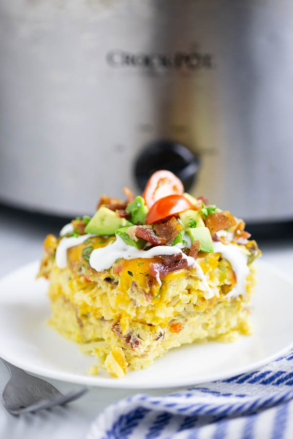 Slice of crockpot breakfast casserole on a white plate with stainless steel crock pot in the background.