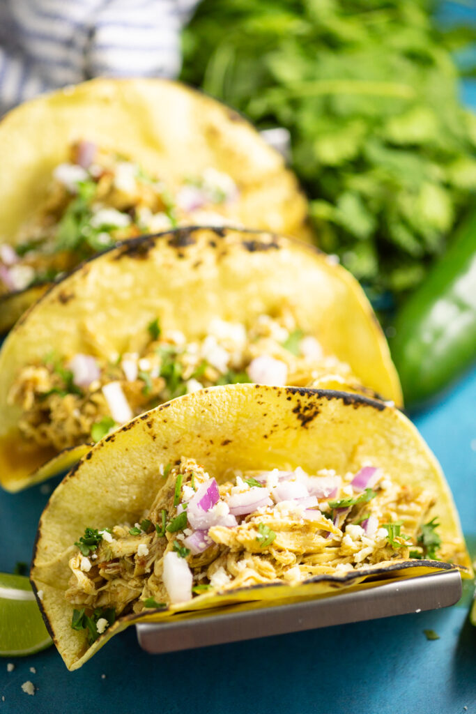 Three crockpot chicken tacos in corn tortillas topped with fresh cilantro and red onion with a fresh jalapeno and fresh cilantro behind.