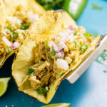 Crockpot chicken taco in corn tortilla topped with fresh cilantro and red onions surrounded by fresh jalapenos and lime wedges.