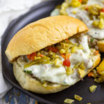 Italian Beef Sandwich topped with provolone and giardiniera on a black plate with a second sandwich sitting behind.