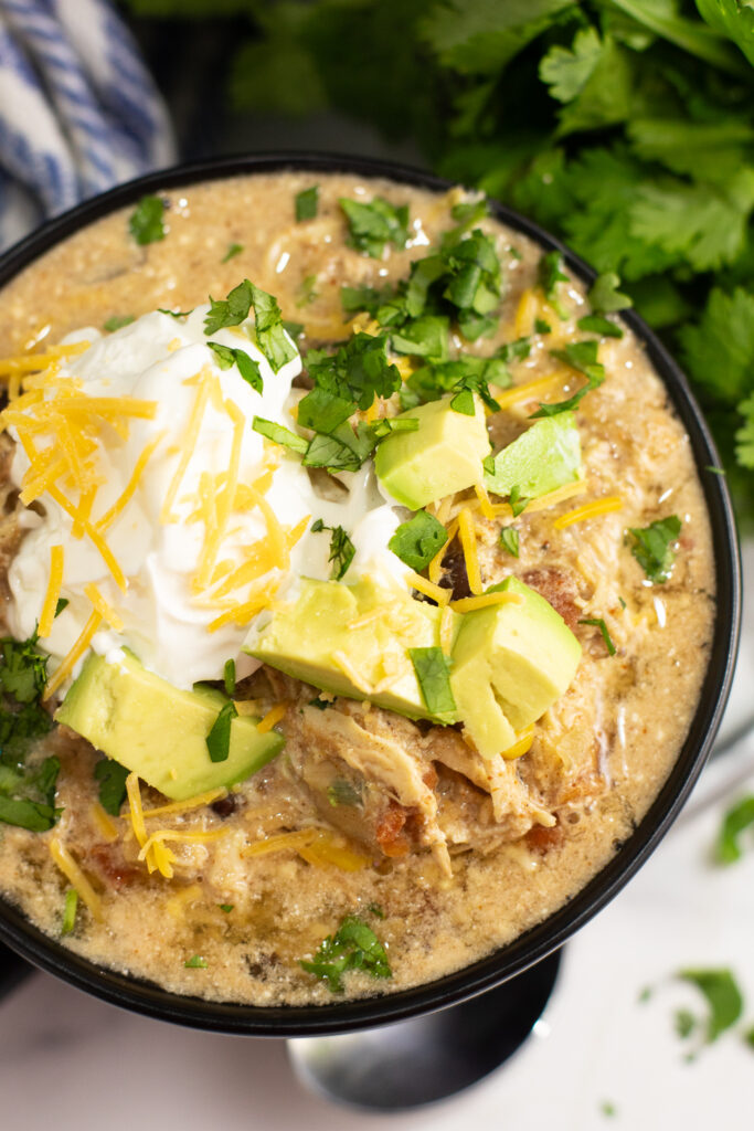 Overhead view of crockpot southwest chicken chili in a black matte bowl with a spoon in it topped with sour cream, shredded cheese, cilantro, and avocado next to a bunch of fresh cilantro and a linen napkin.