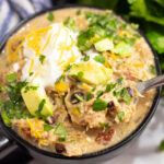 Overhead view of crockpot southwest chicken chili in a black matte bowl with a spoon in it topped with sour cream, shredded cheese, cilantro, and avocado.