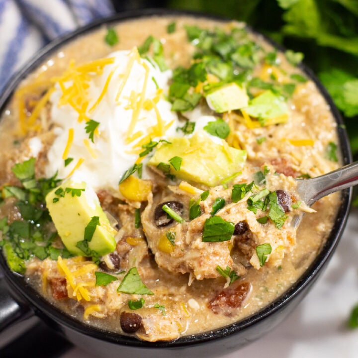 Overhead view of crockpot southwest chicken chili in a black matte bowl with a spoon in it topped with sour cream, shredded cheese, cilantro, and avocado.