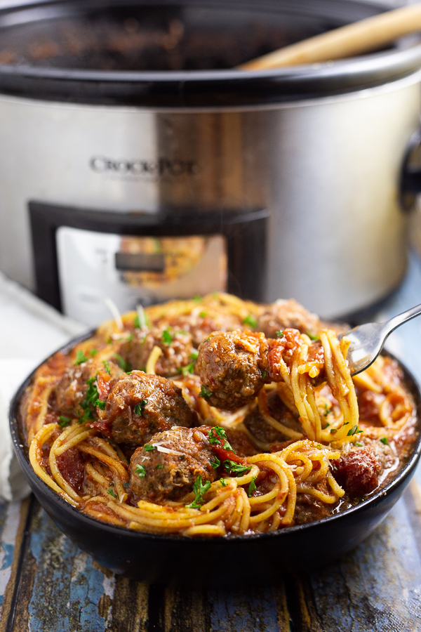 Crock Pot Spaghetti and Meatballs in a shallow bowl with a fork picking some spaghetti and a meatball with a slow cooker in the background