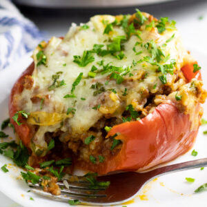 A red stuffed pepper topped with melted cheese and freshly chopped parsley with a bite taken out on a small white plate next to a fork.