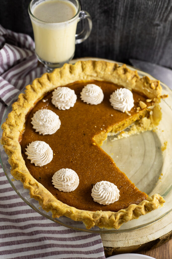 An eggnog pumpkin pie in a glass pie dish topped with piped swirls of whipped cream and a large slice missing. A glass of eggnog and a linen napkin are in the background.