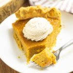 A piece of eggnog pumpkin pie on a small white plate topped with a piped swirl of whipped cream and a fork sitting on the plate with a bite of pie on it.