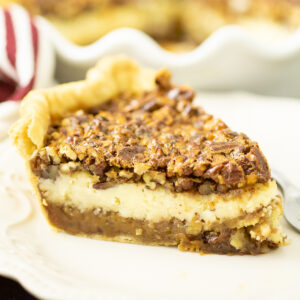 Pecan cheesecake pie on a small white plate with a layer of pie crust, pecan filling, cheesecake, and crunchy pecans.
