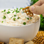 A pretzel dipping into a white matte bowl of ranch beer cheese dip topped with fresh chives with a bunch of parsley behind and crackers and pretzels all around the bottom.