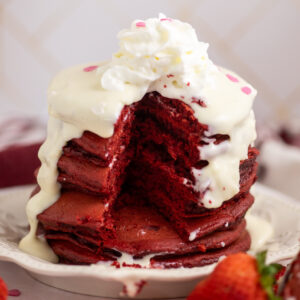 A stack of red velvet cake mix pancakes with a wedge taken out of the center and cream cheese glaze dripping down on a small plate surrounded by fresh strawberries.
