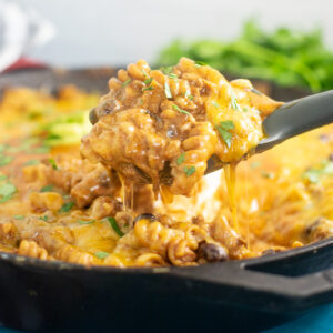A spoon taking a scoop of one pot enchilada pasta from a large cast iron skillet with a bunch of fresh cilantro behind.
