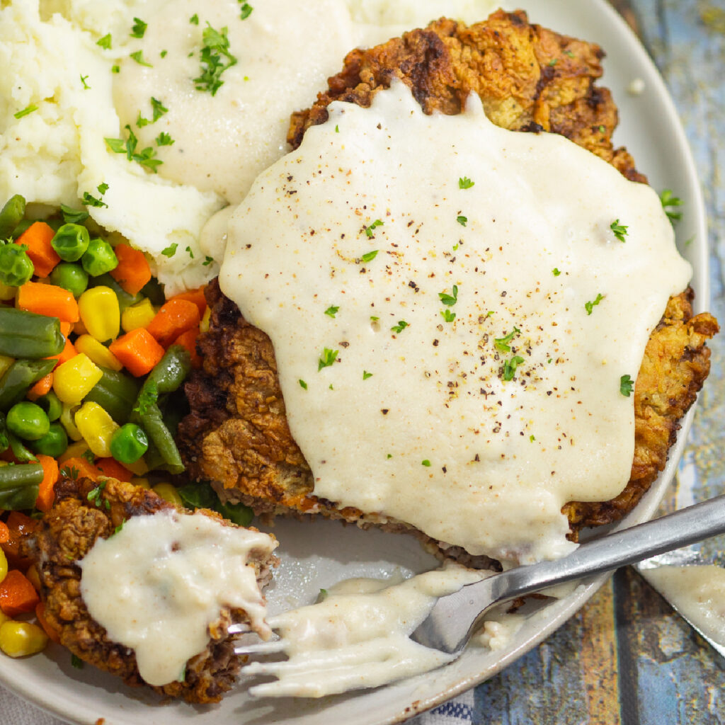 Overhead view of southern chicken fried steak smothered in white gravy and sprinkled with black pepper and parsley on top with a fork cutting a bite off it on a plate with mashed potatoes and mixed vegetables.