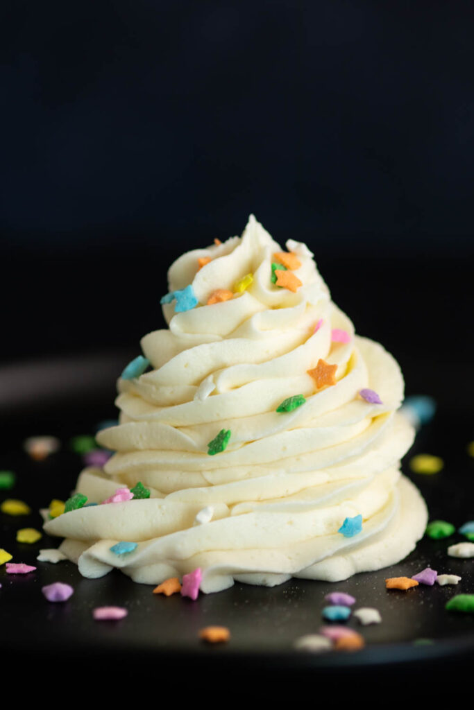 A large swirl of vanilla buttercream frosting on a black background topped with rainbow star sprinkles.