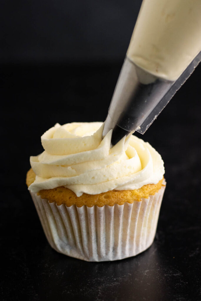 A star decorating tip attached to a piping bag piping vanilla buttercream frosting onto a cupcake on a dark background.