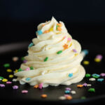 A large swirl of vanilla buttercream frosting on a black background topped with rainbow star sprinkles.