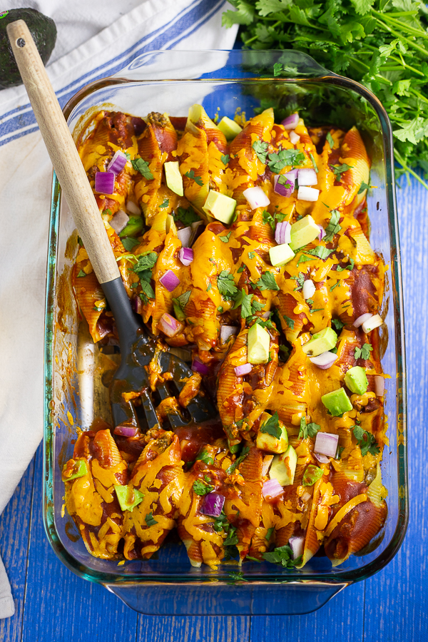 Casserole dish filled with taco stuffed shells topped with fresh avocado, red onion, and cilantro, with a wooden spoon in the middle