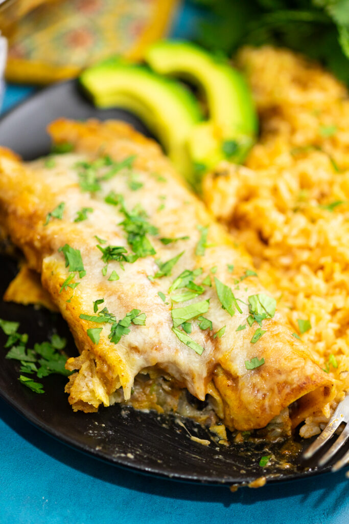 Two black bean enchiladas on a plate with a bite missing topped with fresh cilantro and next to avocado slices and Spanish rice.