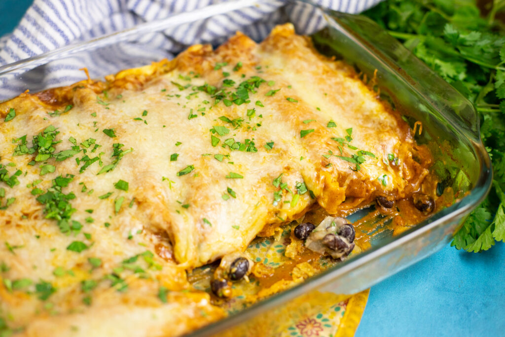 Black Bean enchiladas in a glass baking dish topped with red sauce, melted cheese, and fresh cilantro. The dish is on a bright blue background next to a blue and white striped linen napkin a bunch of fresh cilantro.