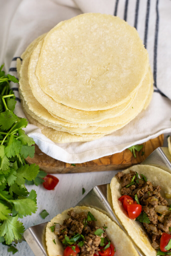 A stack of homemade corn tortillas on top of a striped linen napkin next to two ground beef tacos with cilantro and tomatoes.