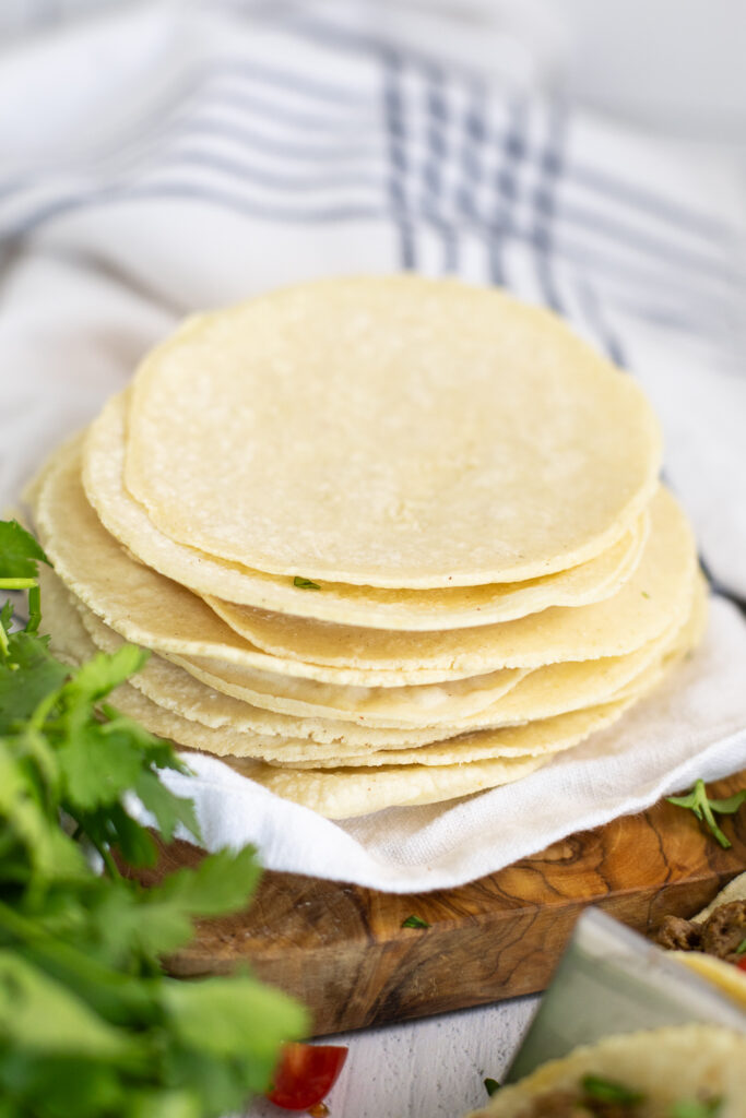 A stack of homemade corn tortillas sitting on top of a wooden cutting board next to a bunch of fresh cilantro.