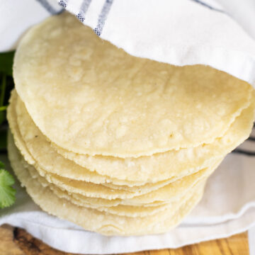 A stack of homemade corn tortillas wrapped in a white linen napkin on a wooden cutting board.